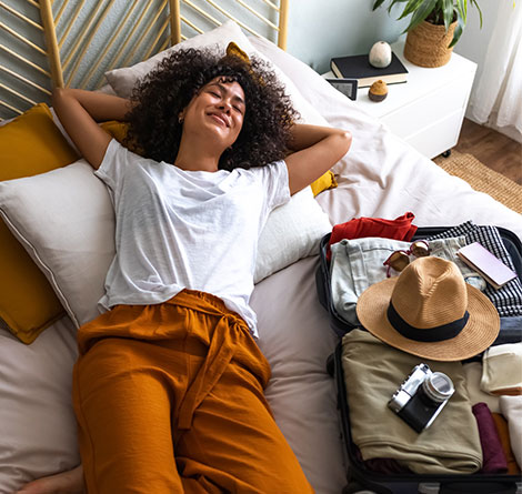 A woman laying on her bed, next to a packed travel bag.
